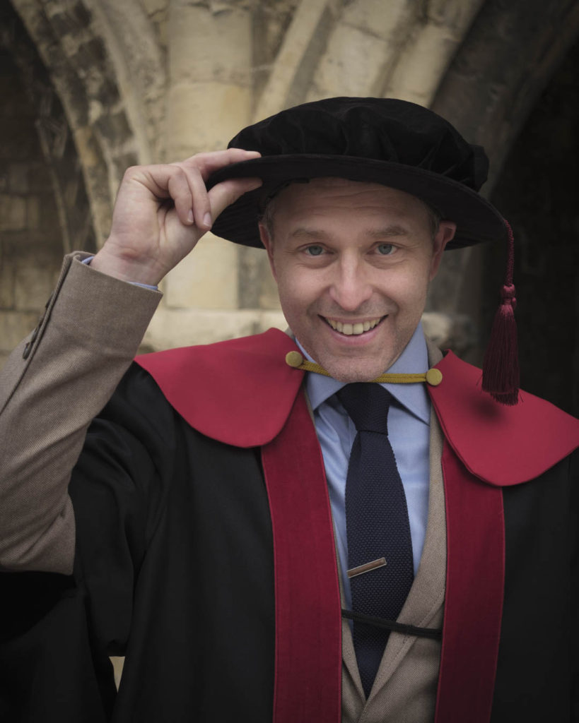 Liam Creighton raises the brim of the PhD bonnet he rented for the day of his graduation. He wears the doctorate gown of the University of Kent: black robes with red trim, red hood with a yellow flash and a black bonnet with a deep red tassel.